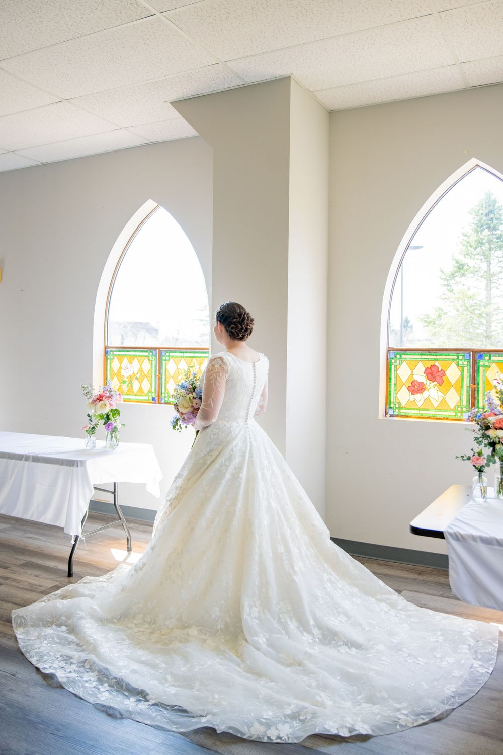 Bride in stunning lace ball gown with long train stands before stained glass chapel windows holding bouquet.