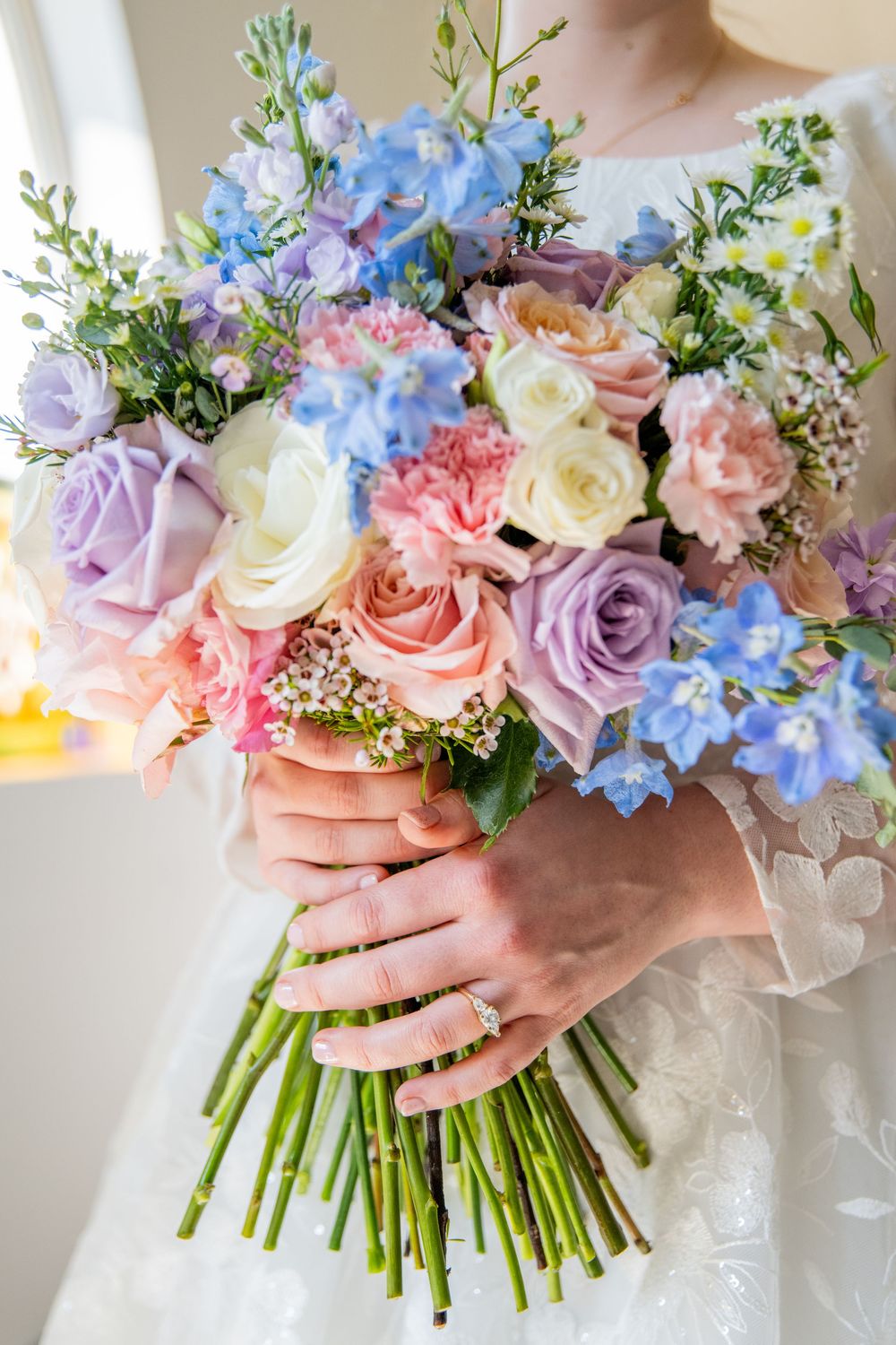 Bride holding a colorful wedding bouquet with pink, purple, blue, and white roses and wildflowers, wearing a diamond ring.