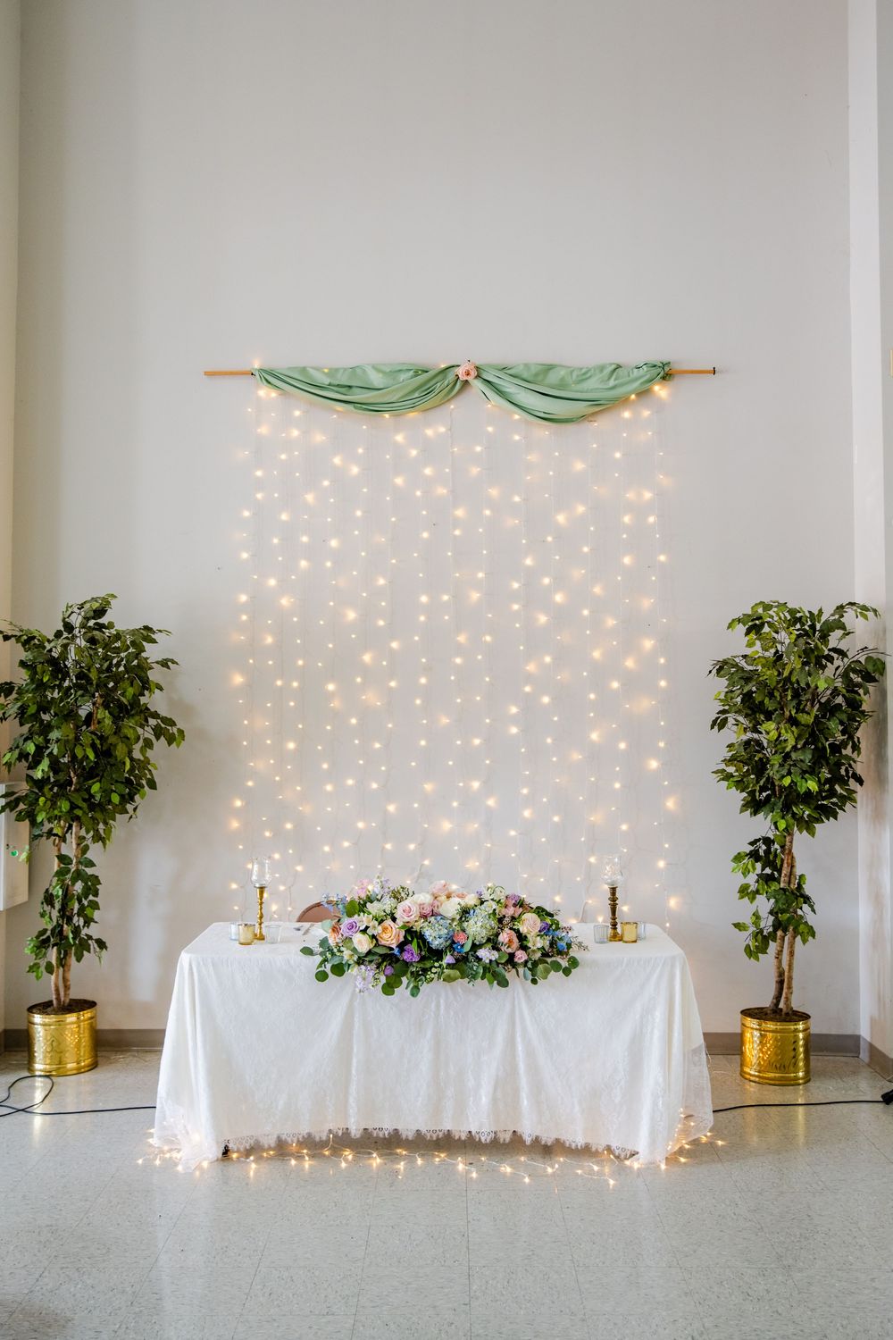 Elegant wedding sweetheart table with floral centerpiece, fairy light backdrop, and sage green draping flanked by potted trees.