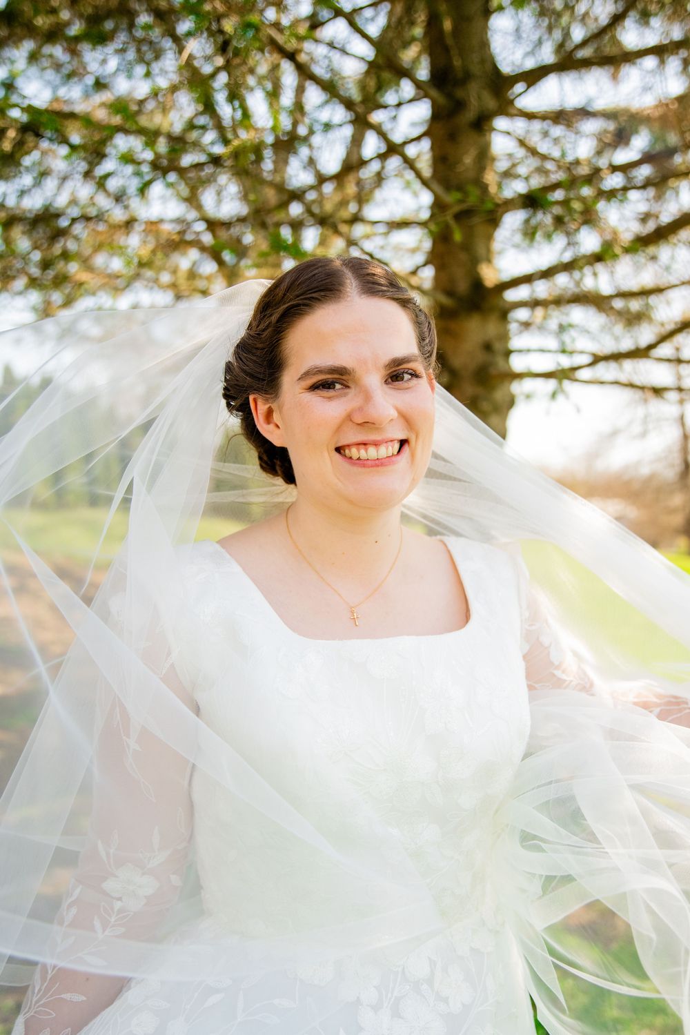 Smiling bride in white lace gown and flowing veil poses outdoors under a tree on her wedding day.