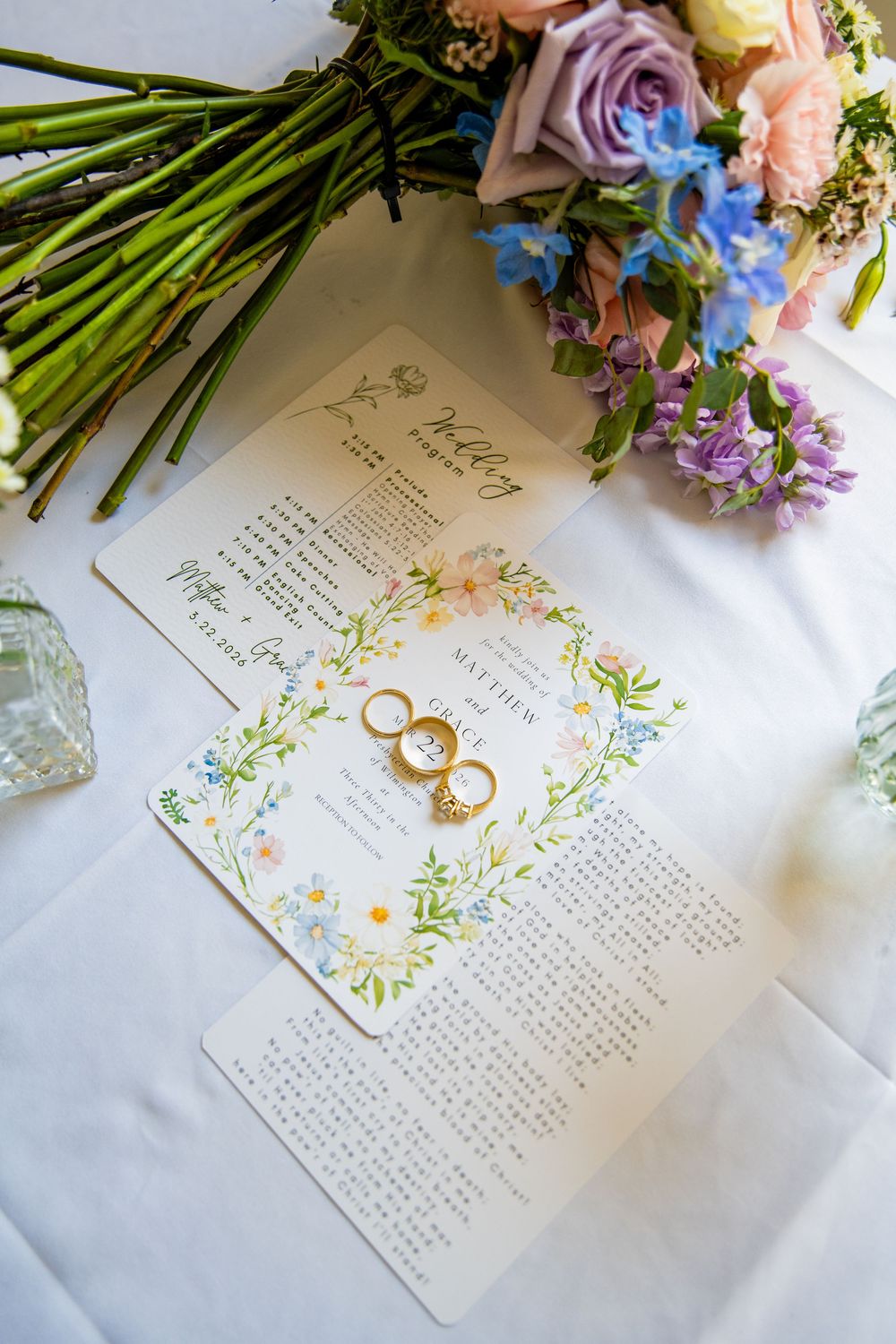 Wedding program and rings displayed on a table with colorful floral bouquets of pink, purple, and blue flowers.