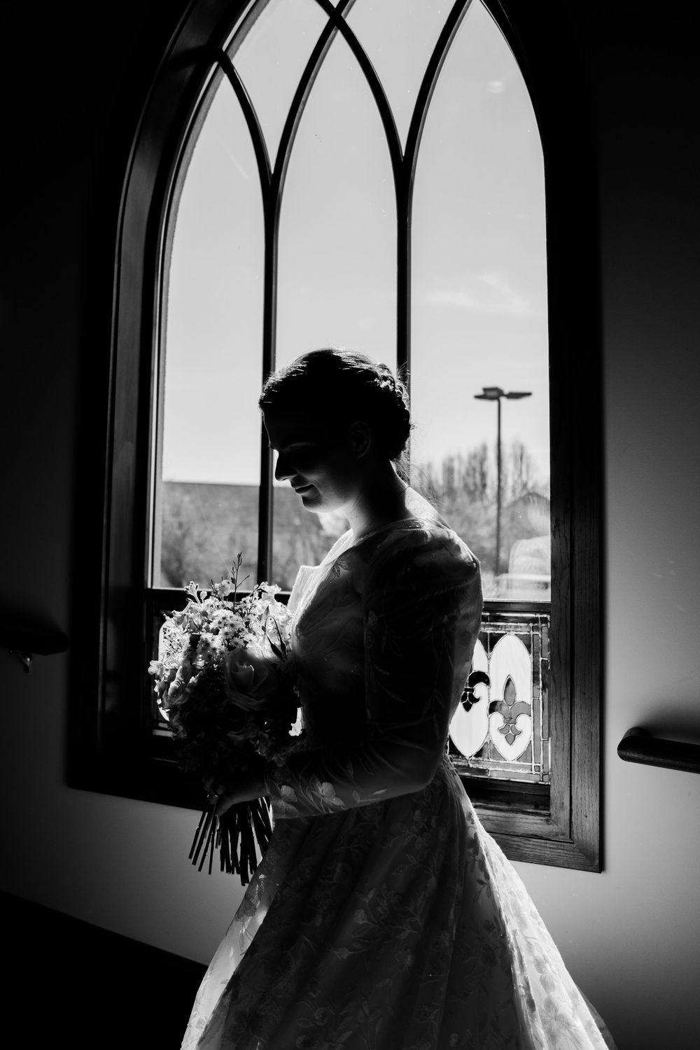 Black and white silhouette of a bride holding a bouquet standing before an arched church window on her wedding day.