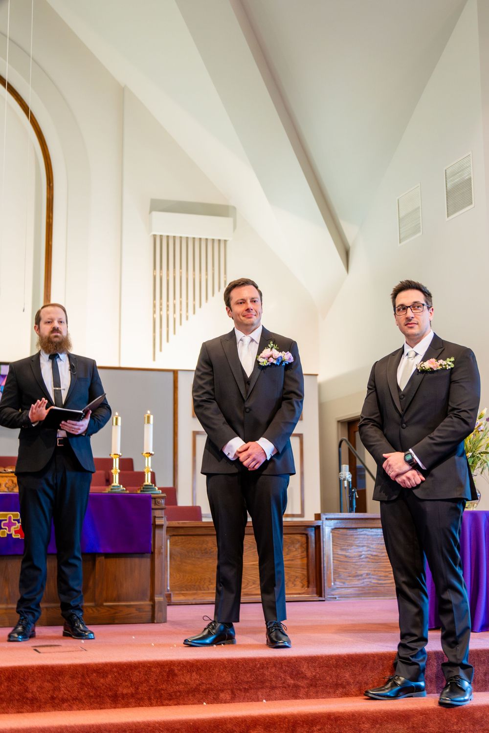 Groom and groomsmen in black suits stand at church altar during wedding ceremony with officiant.