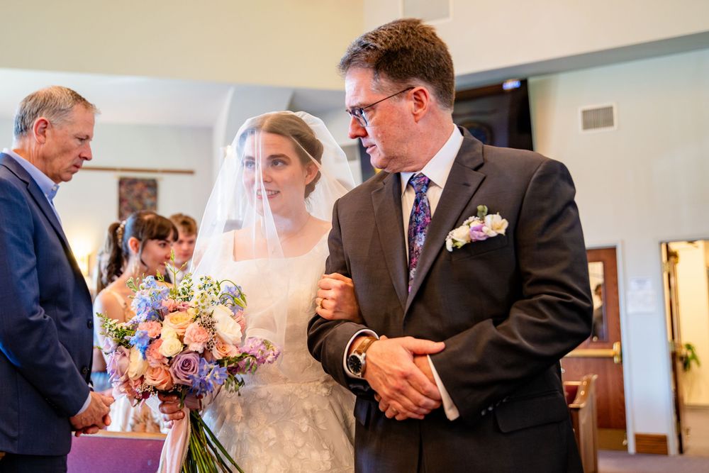 Father escorts bride in white gown and veil down church aisle holding colorful wedding bouquet.