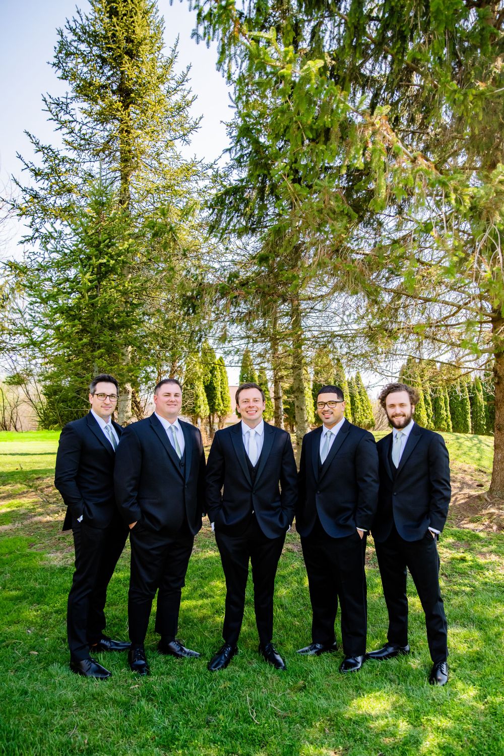 Five groomsmen in matching black suits posing outdoors on green lawn with tall evergreen trees behind them.