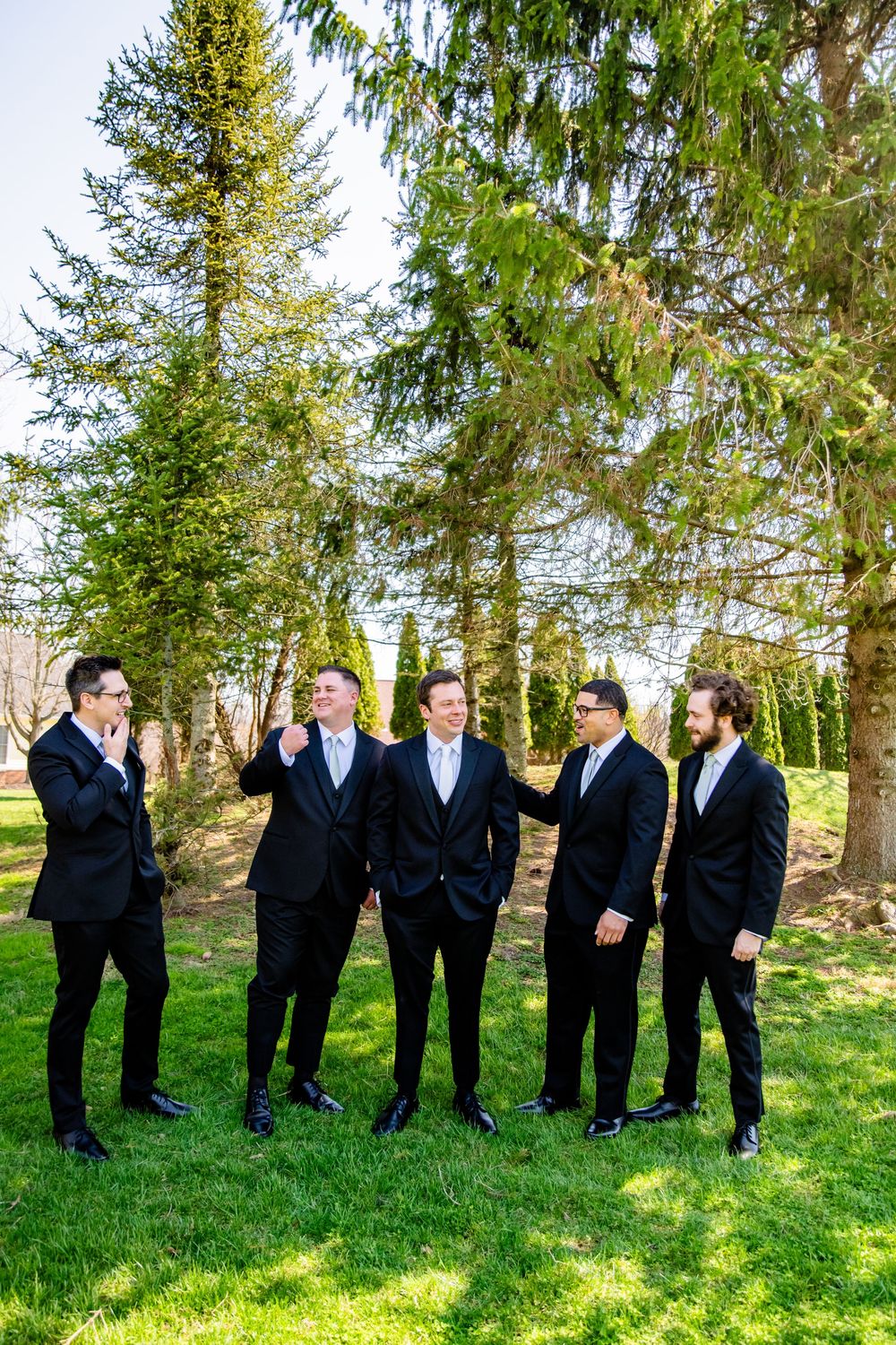 Five groomsmen in black tuxedos laughing together on green grass with tall evergreen trees in background.