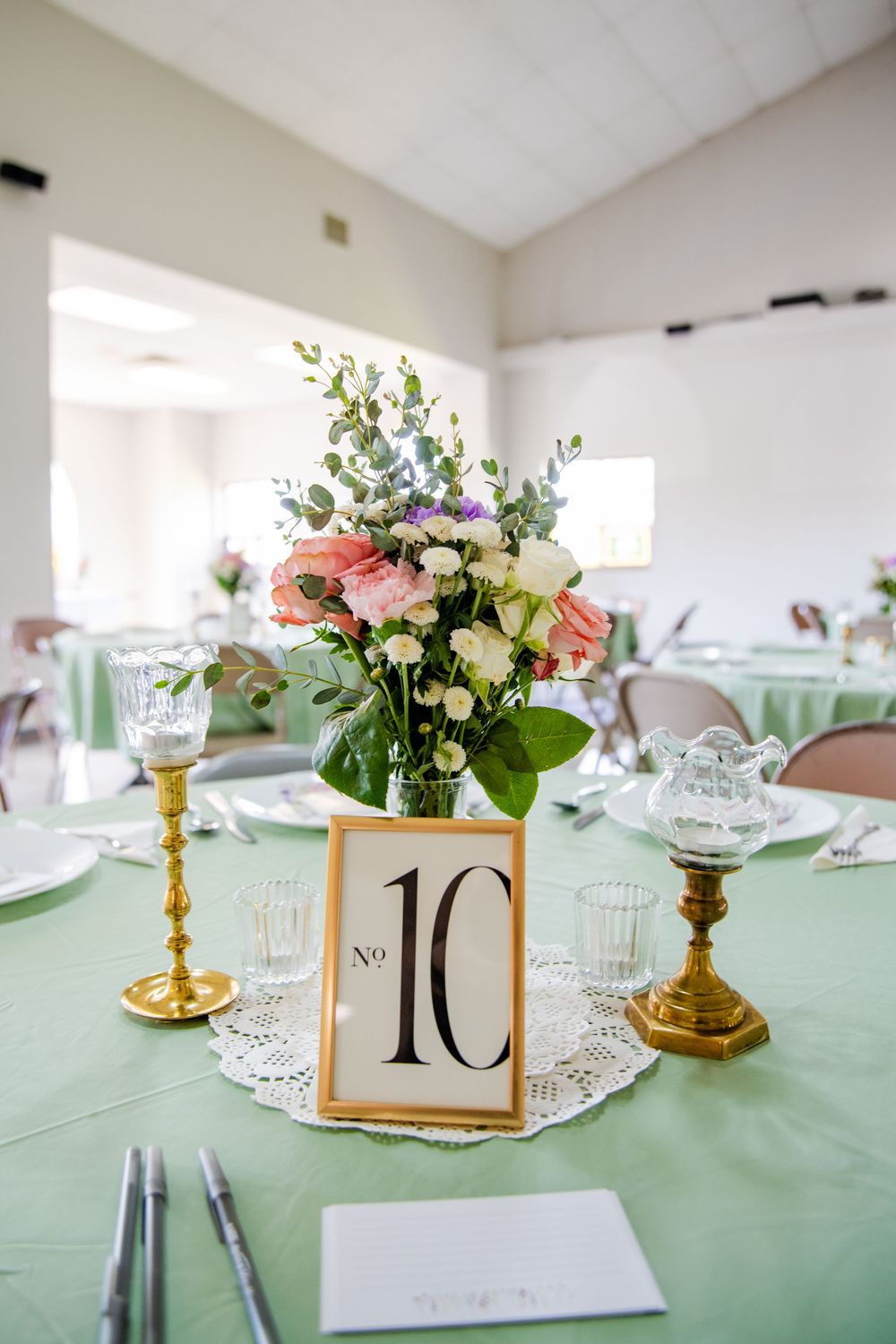 Elegant wedding reception table with mint green linens, gold candlesticks, pink floral centerpiece, and table number 10 sign.