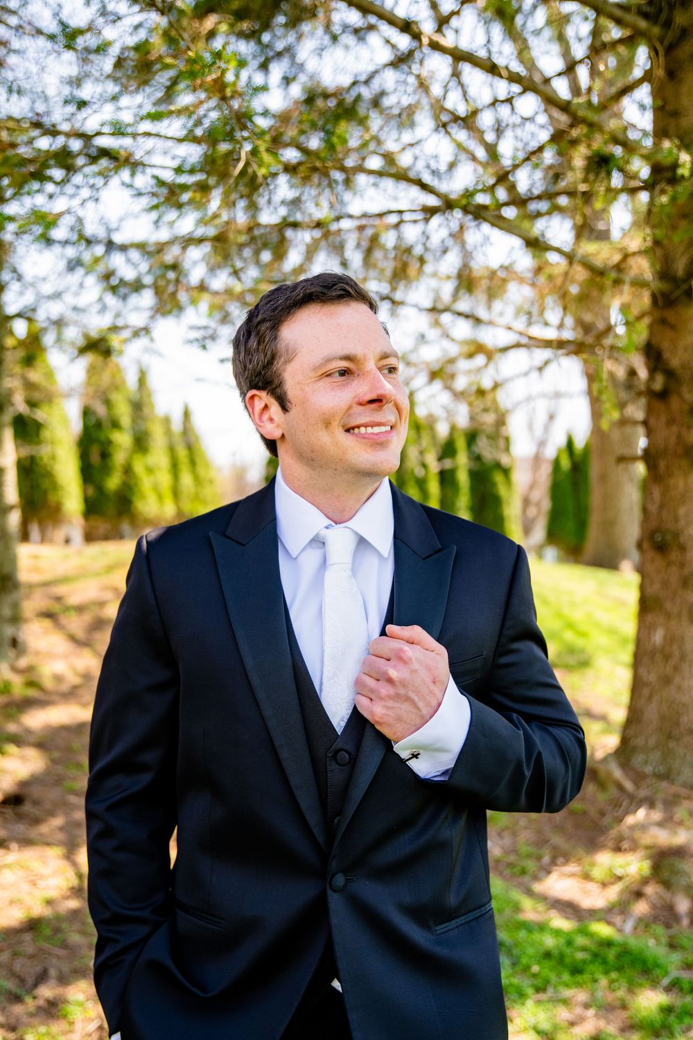 Smiling groom in black suit and white tie standing outdoors among green trees on wedding day.