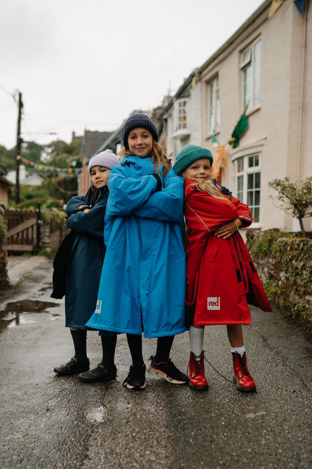 Three children posing confidently in navy, blue, and red waterproof coats on a rainy village street.