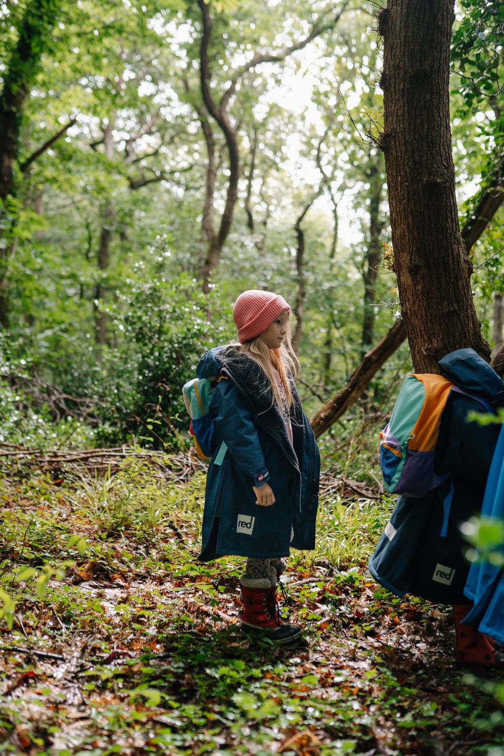 A young girl in a teal coat and pink hat explores a lush green forest trail with a backpack.