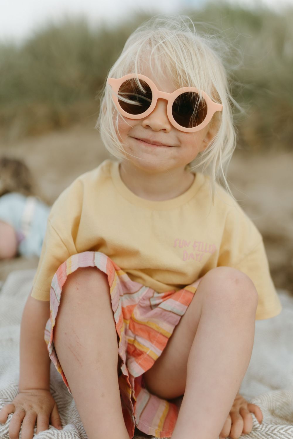 Smiling blonde child wearing peach sunglasses, yellow tee, and plaid shorts sitting outdoors.