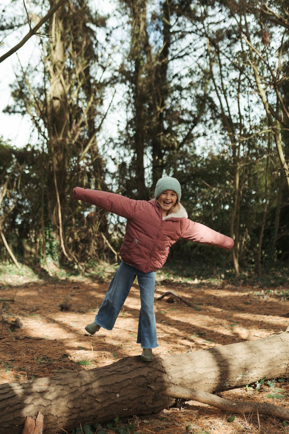 Girl balancing on a fallen log in a forest, wearing a pink puffer jacket and sage green beanie hat.