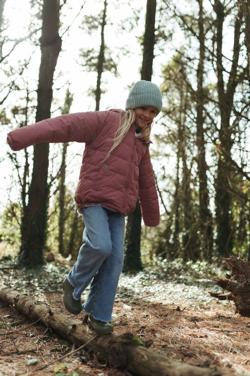 A young girl in a pink puffer jacket and grey beanie balances on a fallen log in a woodland forest.