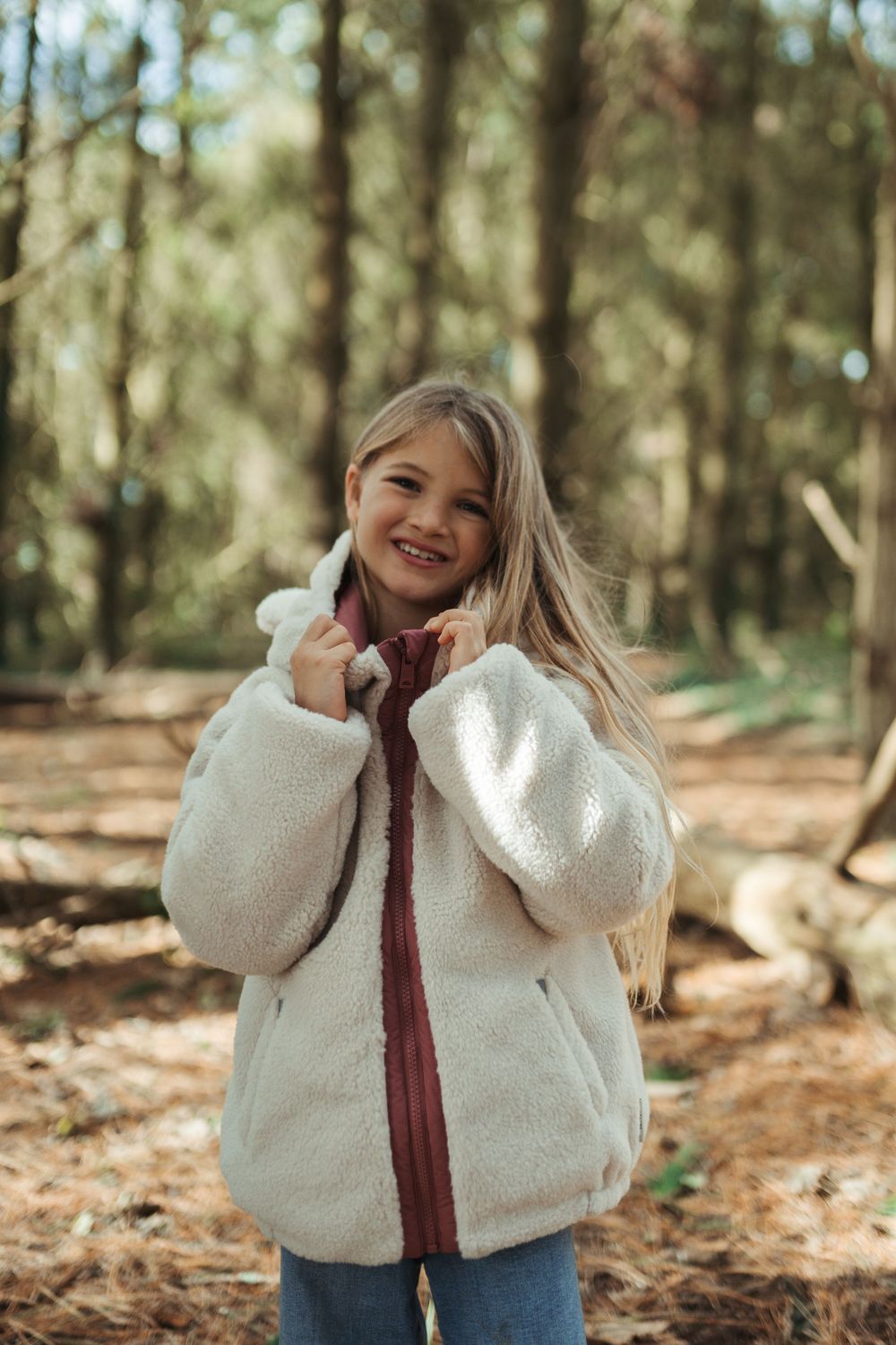 Smiling girl in white fluffy teddy coat standing in a sunlit forest.