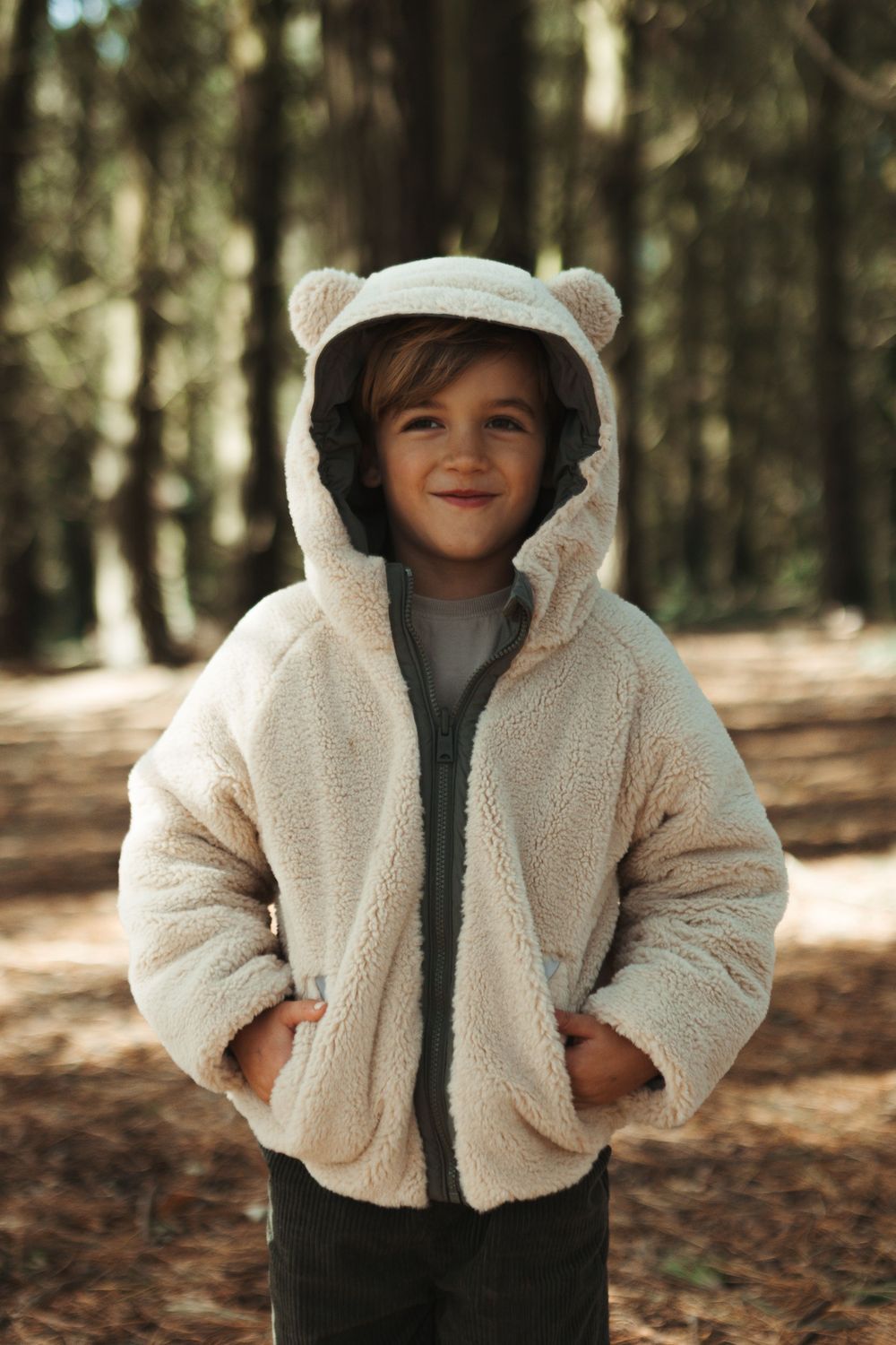 Smiling child in a cream sherpa bear ear hoodie jacket standing in a sunlit forest.