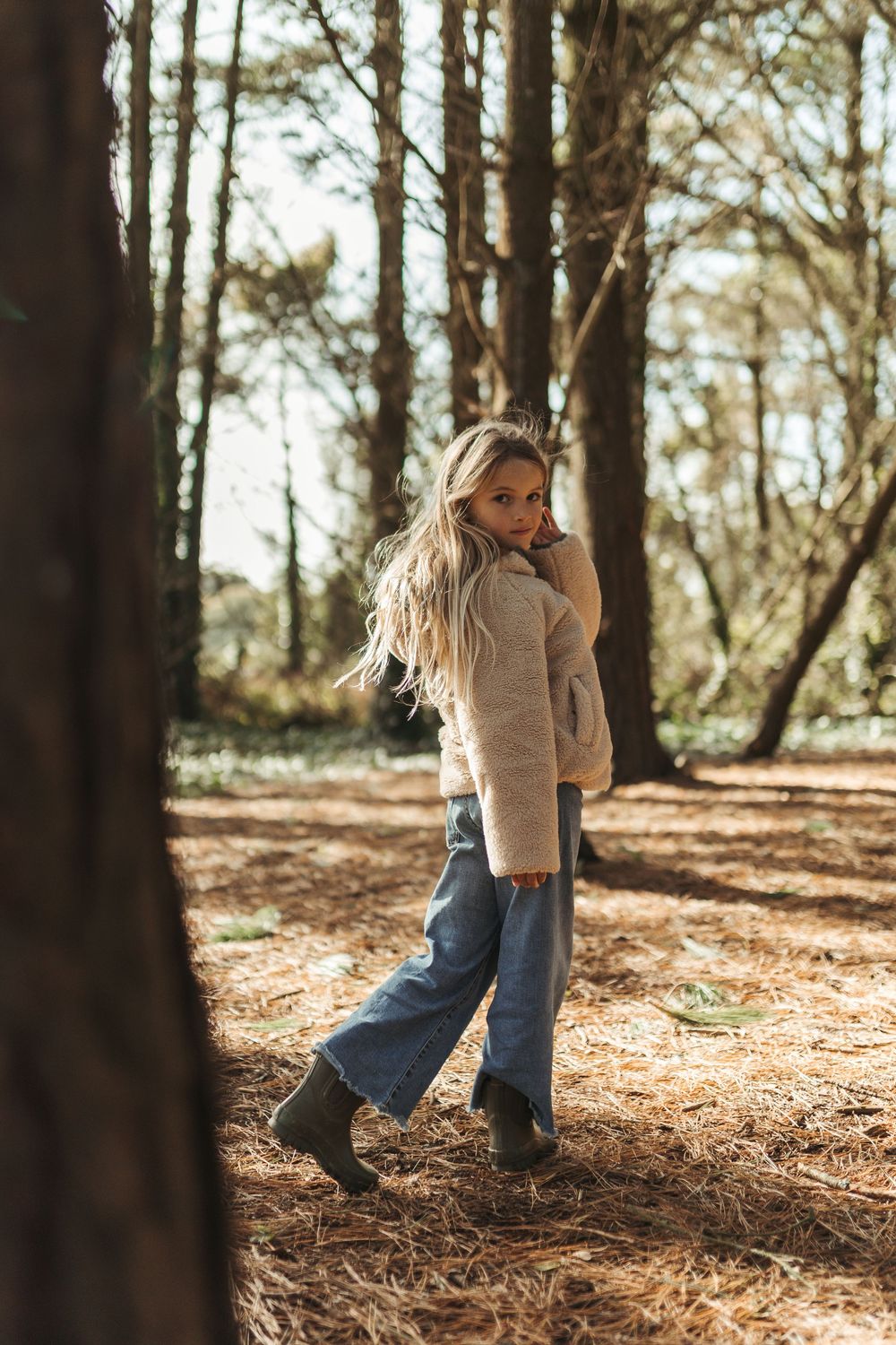 Young woman with long blonde hair walks through a sunlit pine forest wearing a cream sweater and blue wide-leg jeans.