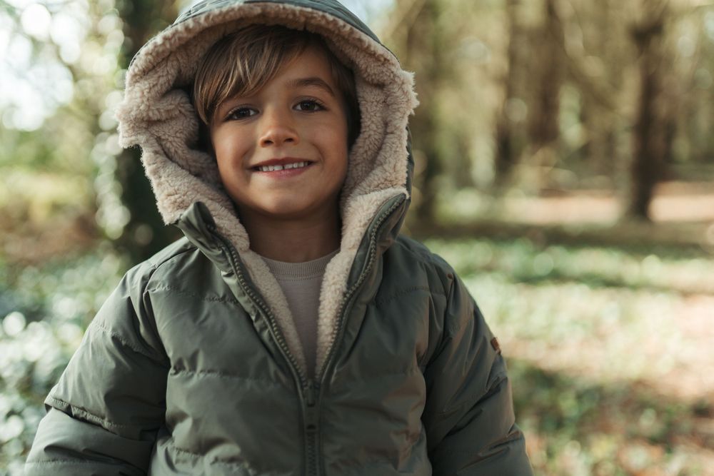 Happy boy in green hooded puffer jacket smiling outdoors in a forest.