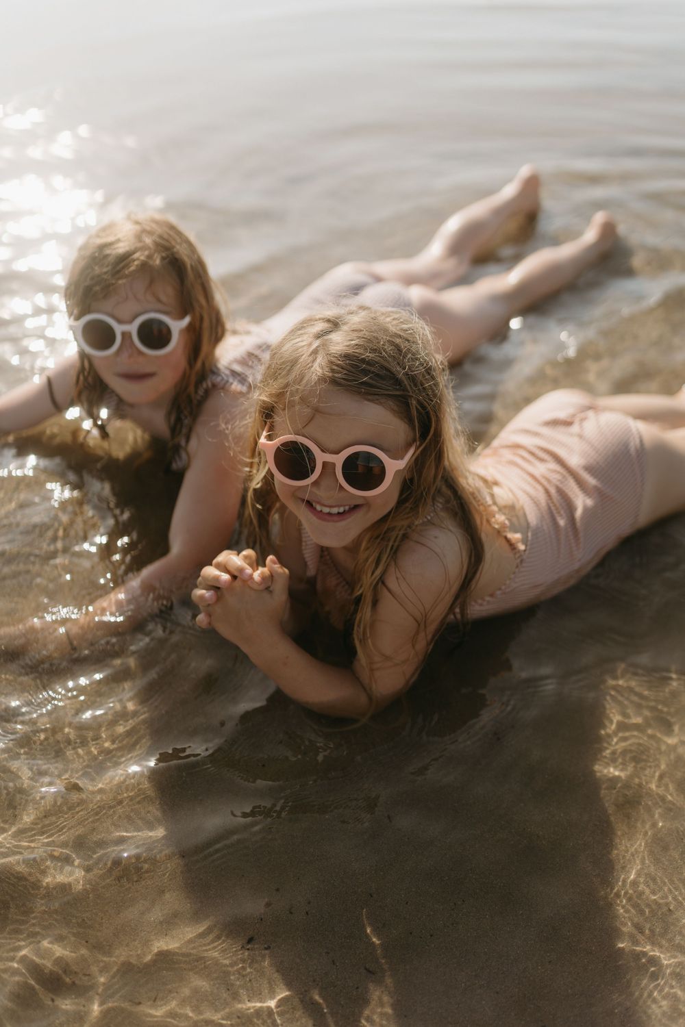 Two young girls wearing sunglasses lying in shallow water at the beach, smiling and enjoying summer.