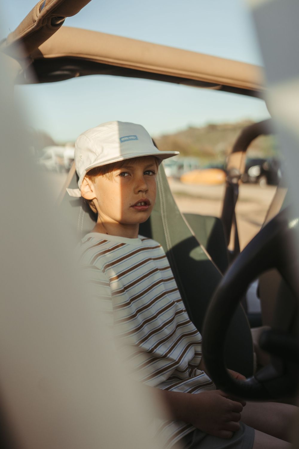 Young woman in striped shirt and white cap sitting inside a convertible car on a sunny day.