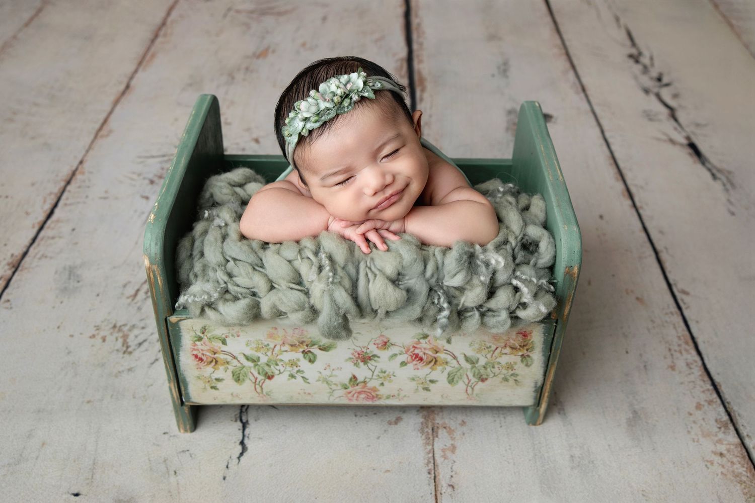 Smiling newborn baby in a tiny green wooden bed with floral details, wearing a delicate flower headband.