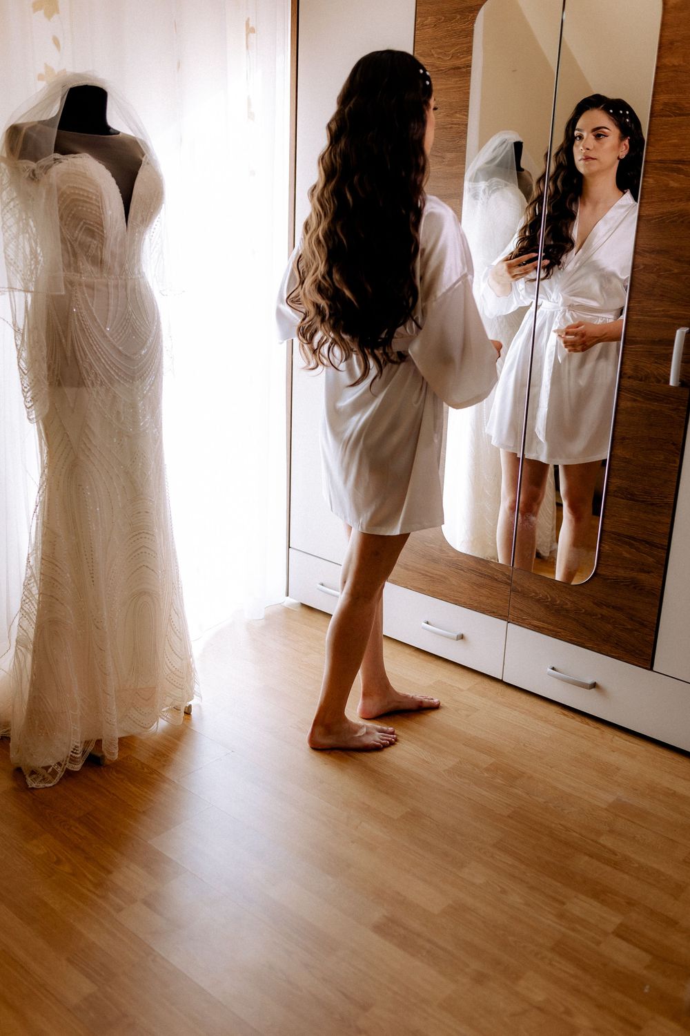 Bride in white robe admiring her lace wedding dress on mannequin while looking in mirror.