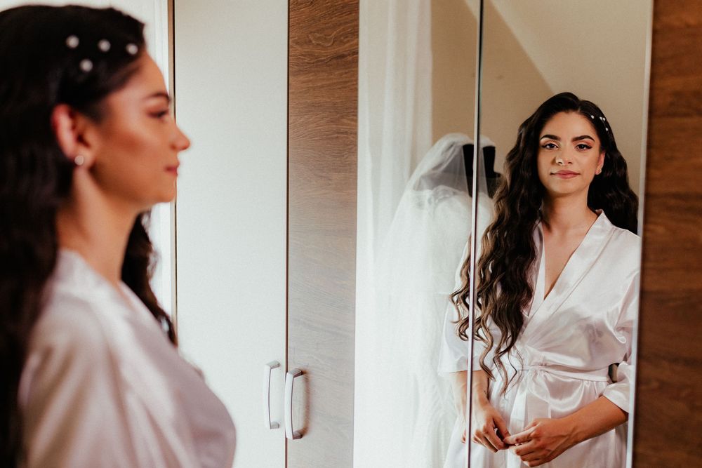 A beautiful bride in a white robe admires her wedding look in a mirror with her gown visible behind her.