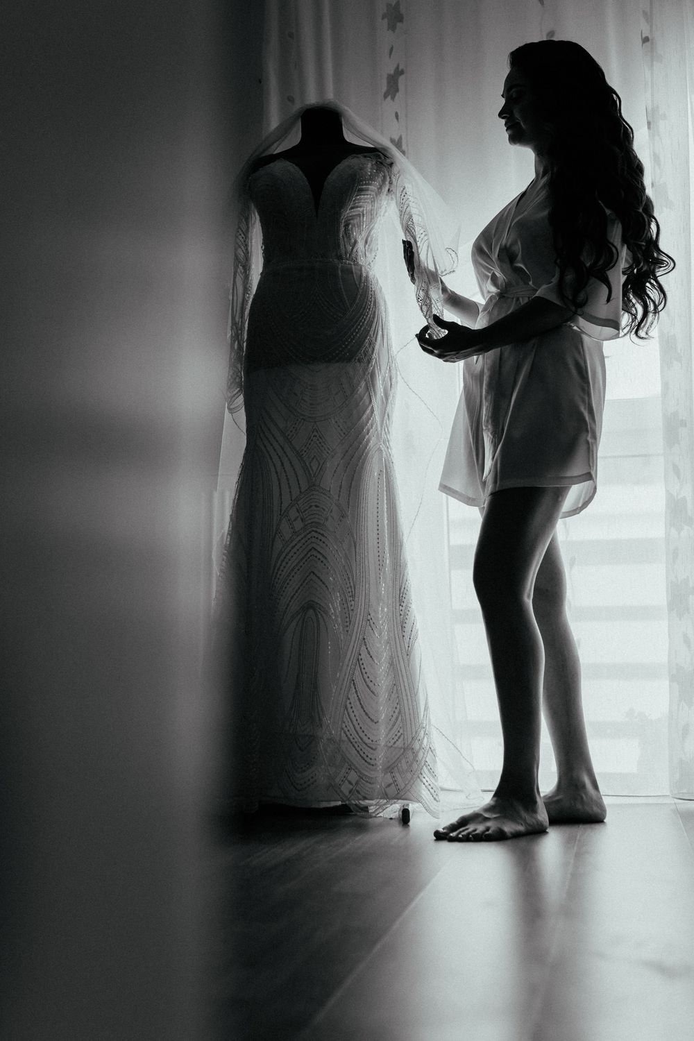 Black and white photo of a bride in a robe admiring her wedding dress on a mannequin by a bright window.