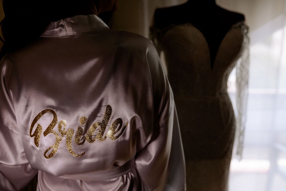 Bride in satin robe with gold glitter script stands before elegant wedding gown on mannequin in soft light.