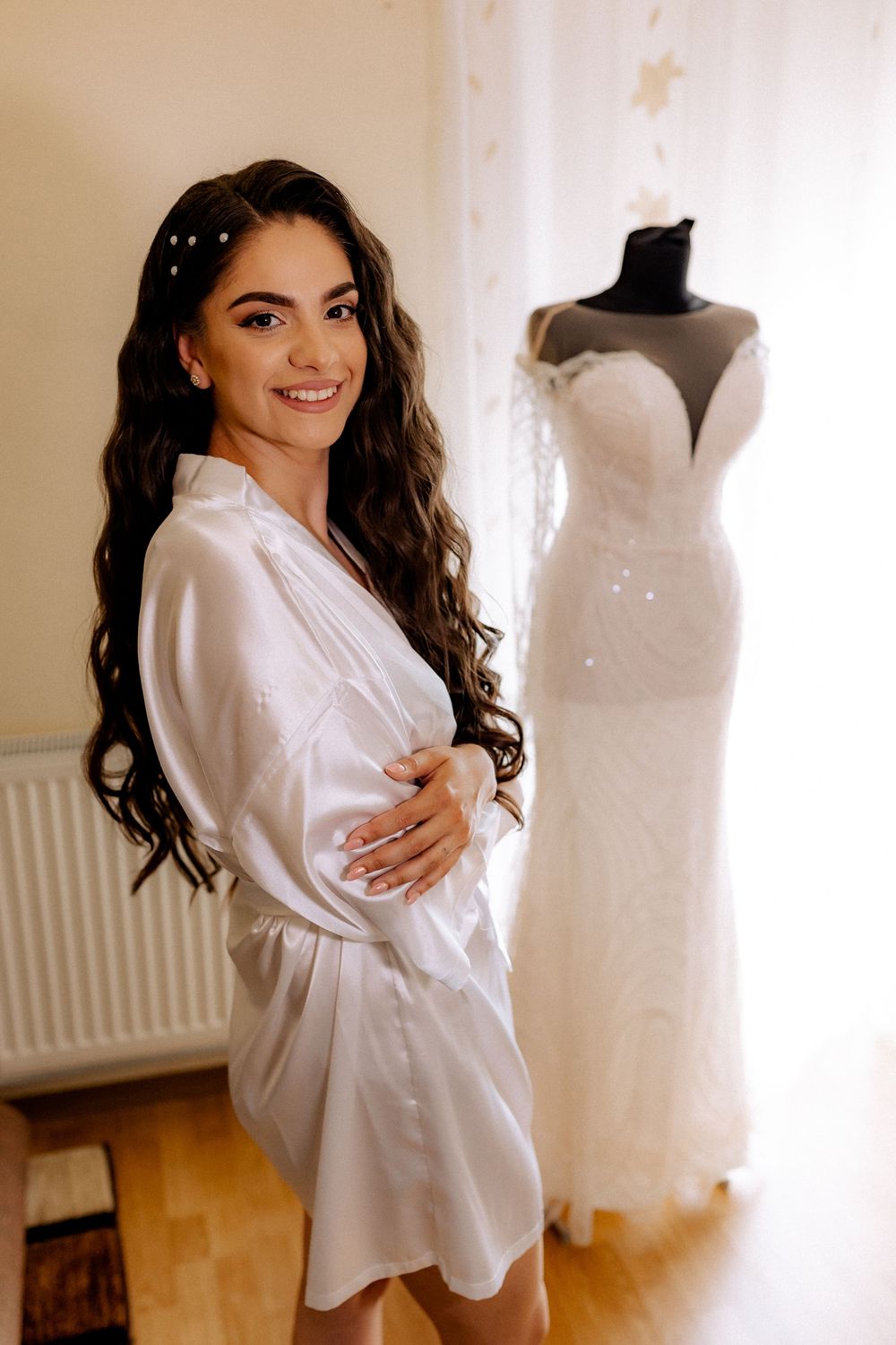 Smiling bride in white satin robe stands before wedding gown on mannequin during bridal preparation.