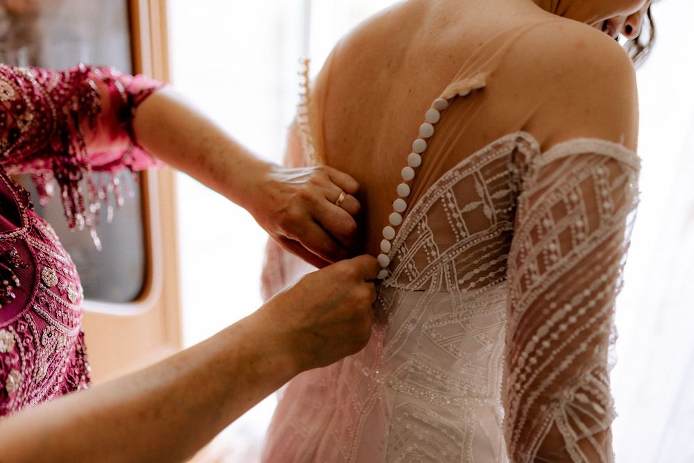 Helper fastening pearl buttons on back of lace wedding dress bridal preparation.