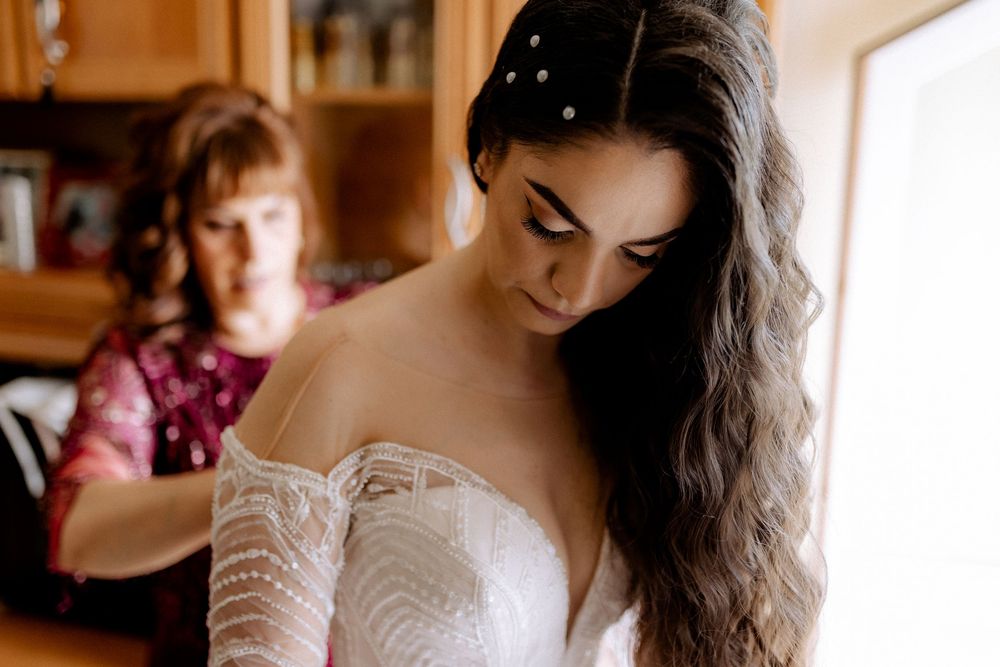 Bride in lace off-shoulder gown with pearl hair accessory being helped to dress.