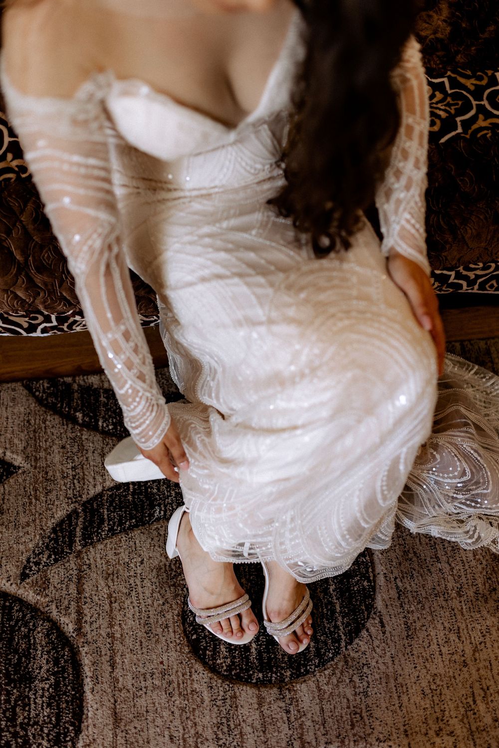 Bride in beaded long-sleeve wedding gown putting on white heeled sandals overhead view.
