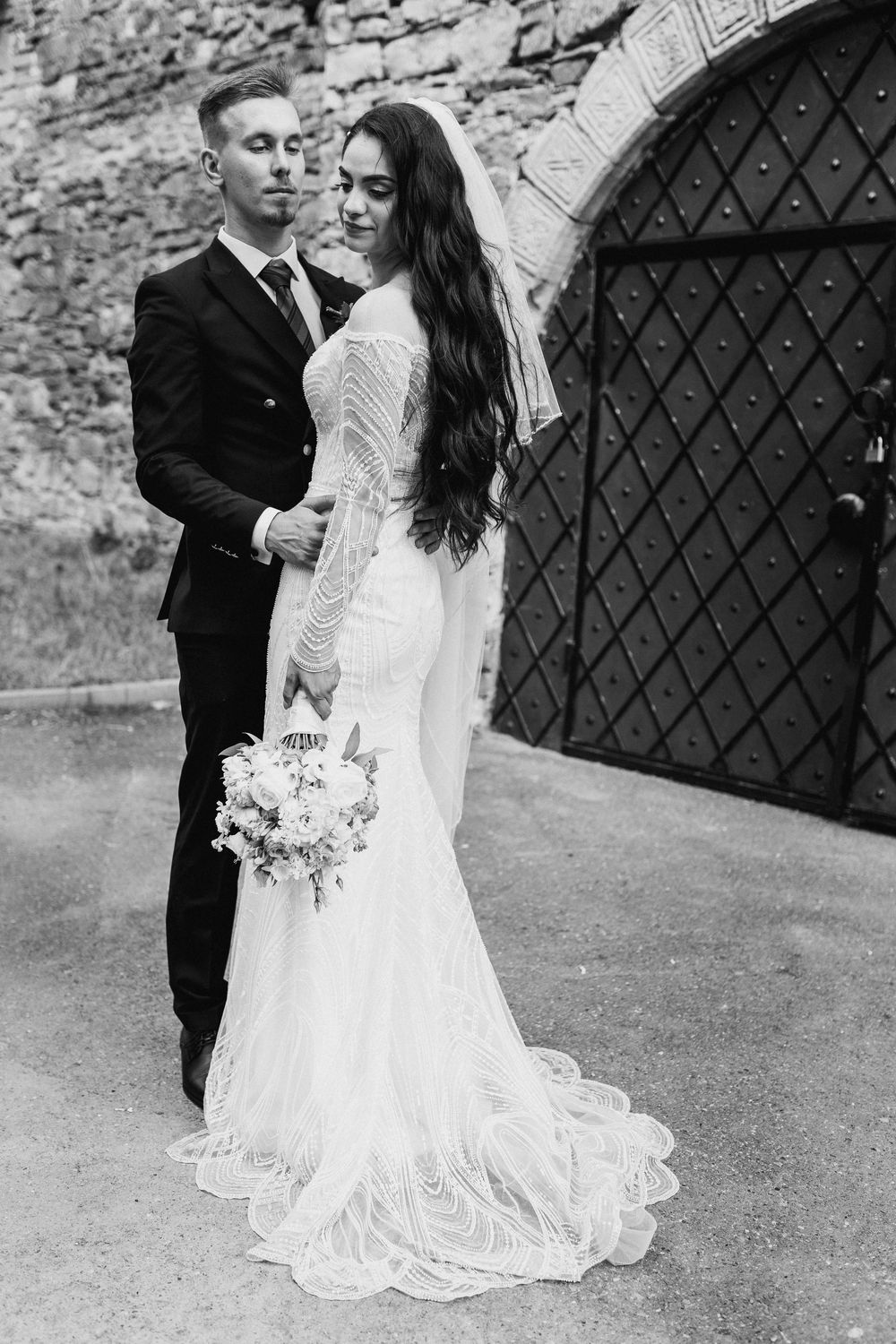 Black and white photo of bride and groom embracing near stone wall and ornate door.