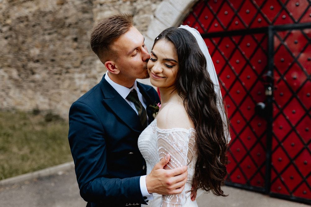 Groom in navy suit kissing brides cheek as she smiles near red decorative doors.