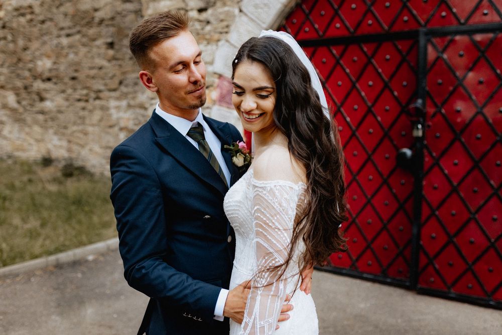 Happy newlyweds embracing outdoors, groom in navy suit, bride in lace dress with veil.