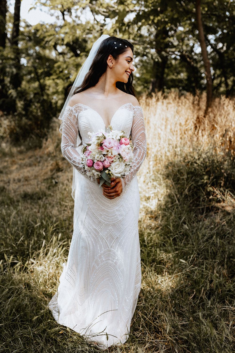 Smiling bride in lace long-sleeve wedding gown holding pink and white bouquet, standing in sunlit outdoor field.