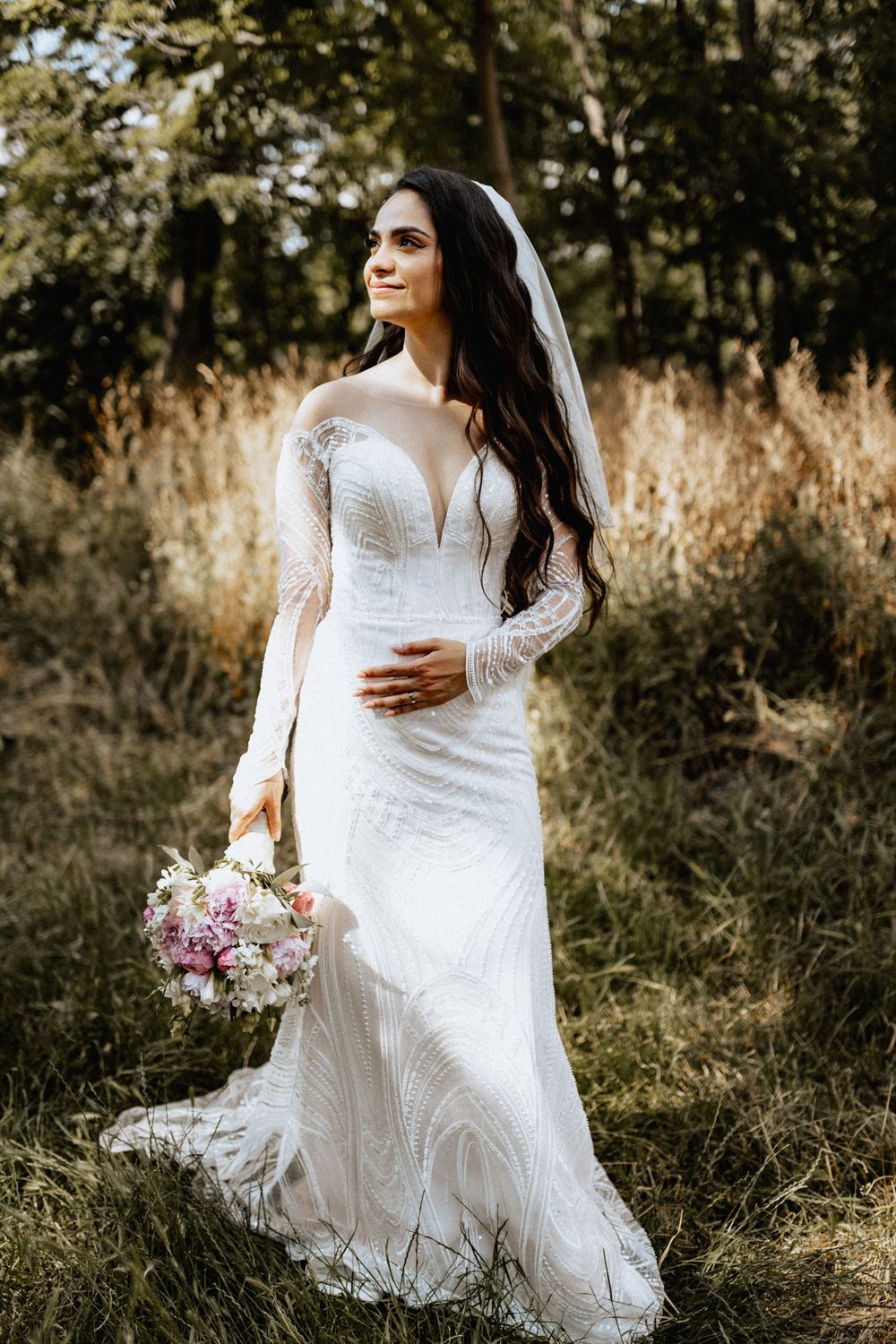 Elegant bride in lace wedding gown holding bouquet while standing in a sunlit field.