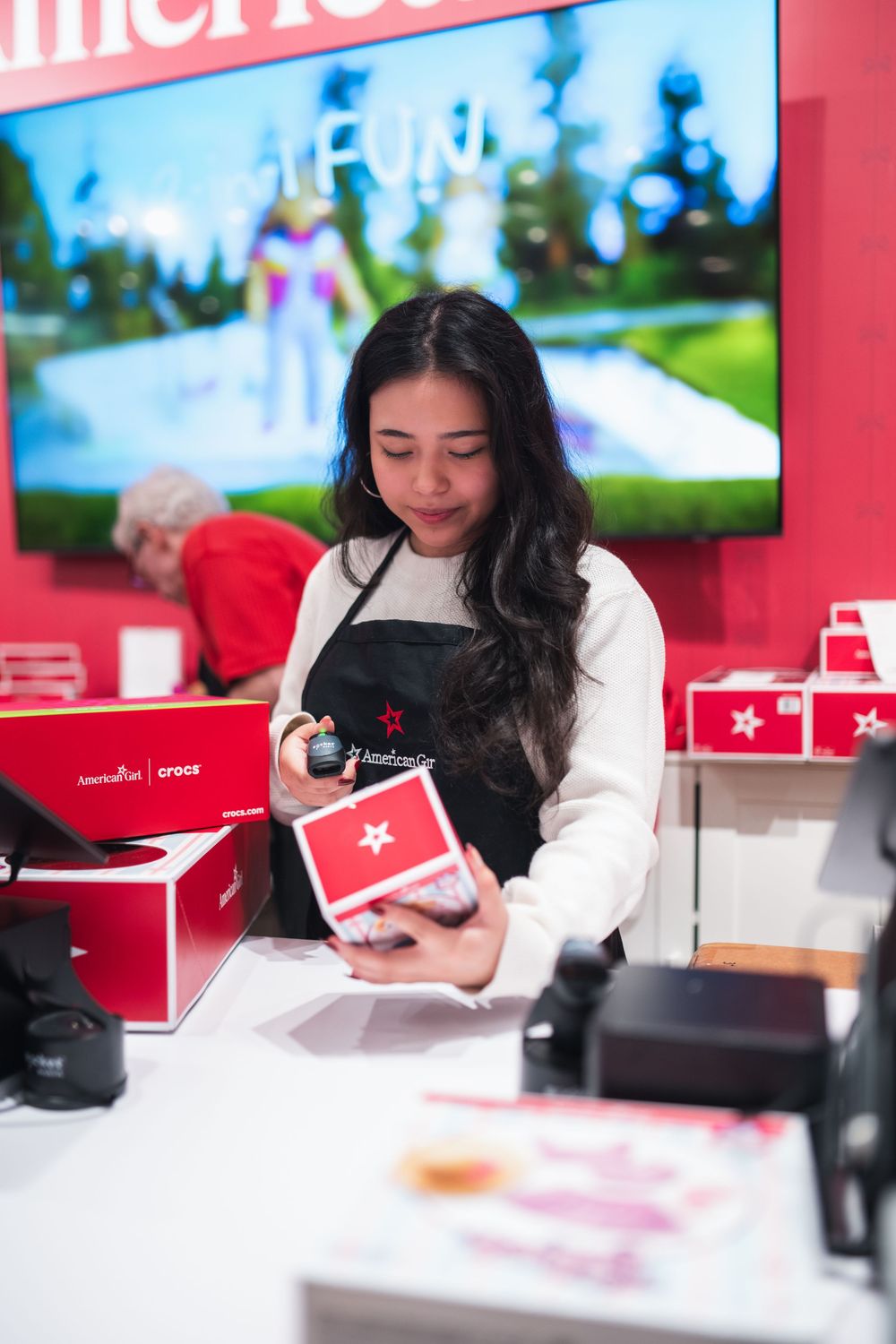 A young woman in a black apron scans a red gift box at an American Girl store checkout counter.