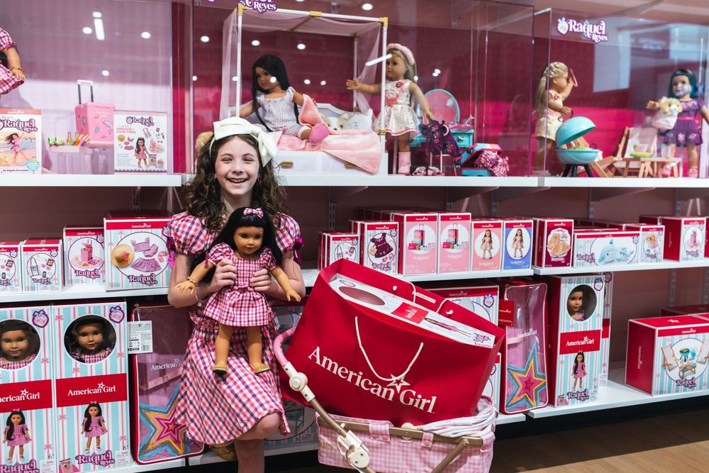 A smiling girl holds an American Girl doll and shopping bag in front of a colorful American Girl store display.