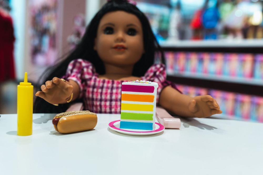 American Girl doll sitting at a table with miniature rainbow cake, hot dog, and mustard bottle toys.