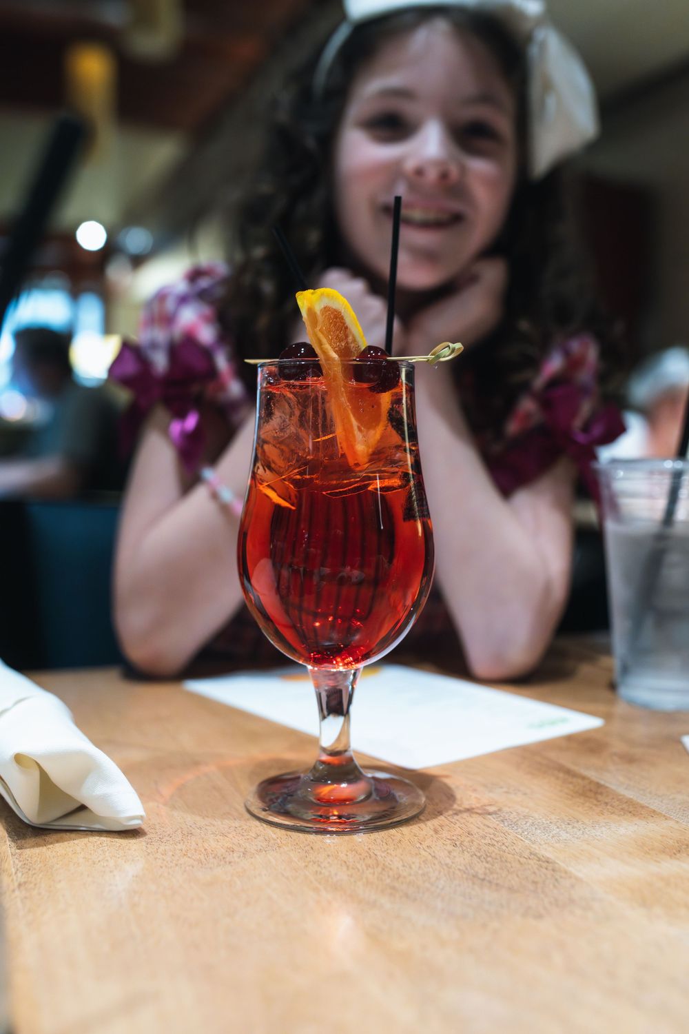A young girl smiles behind a red cocktail garnished with orange slice and cherries at a restaurant table.