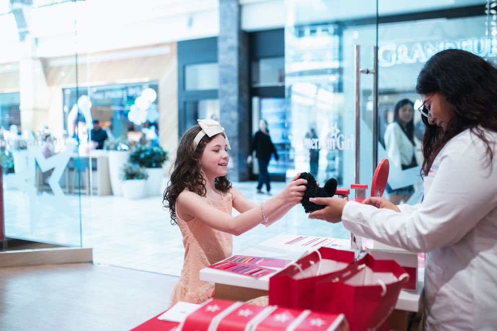 A young girl in a pink dress and tiara receives a gift box from a woman in a white coat at a mall kiosk.