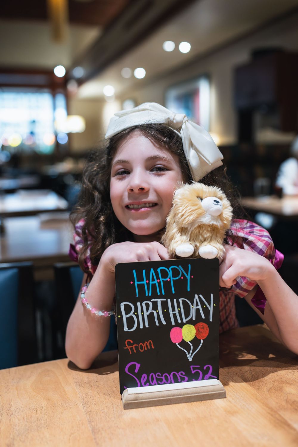 Young girl smiling at Seasons 52 restaurant with a stuffed animal and Happy Birthday chalkboard sign.
