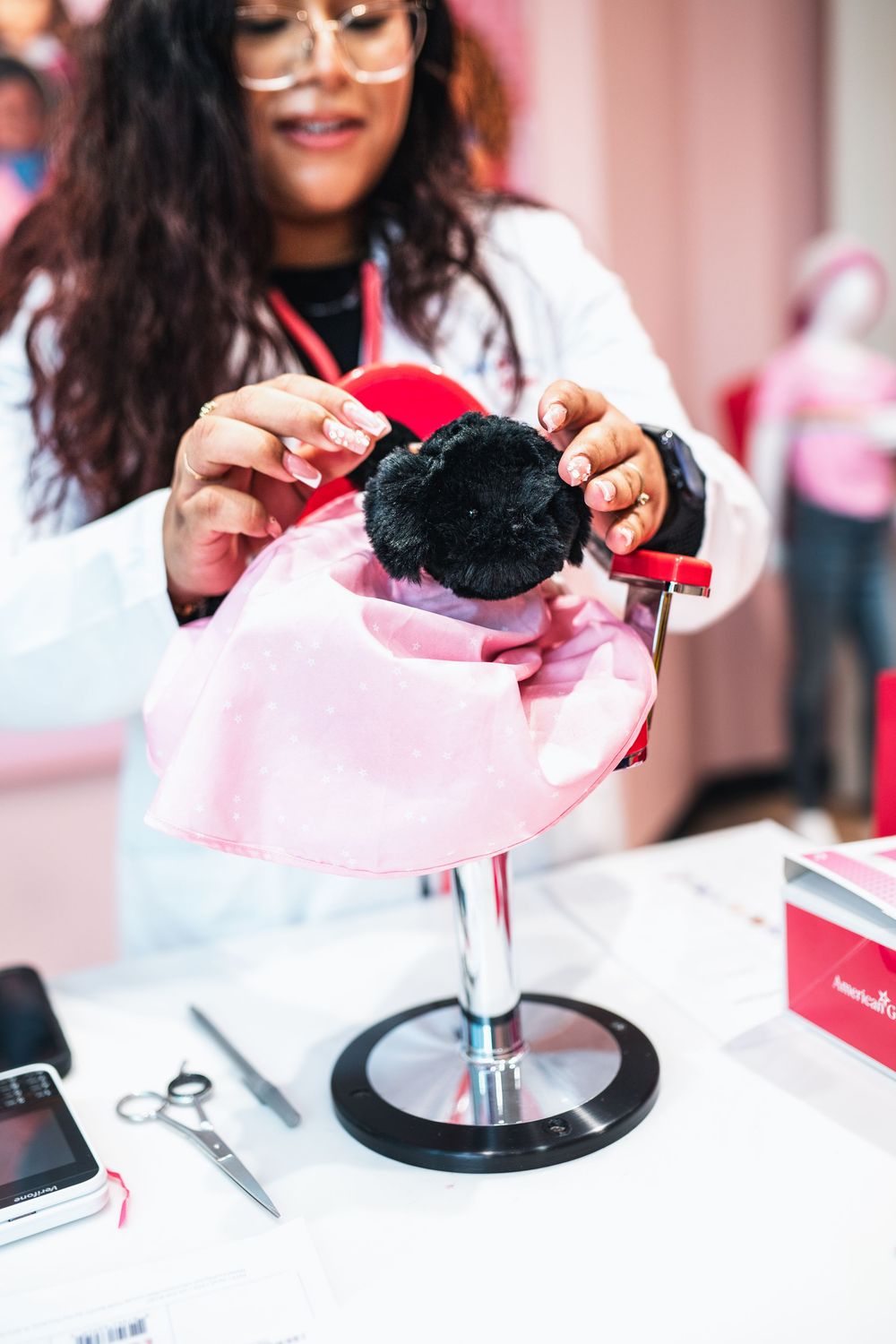 A woman in a white coat styles a doll's hair on a miniature salon chair at an American Girl store.