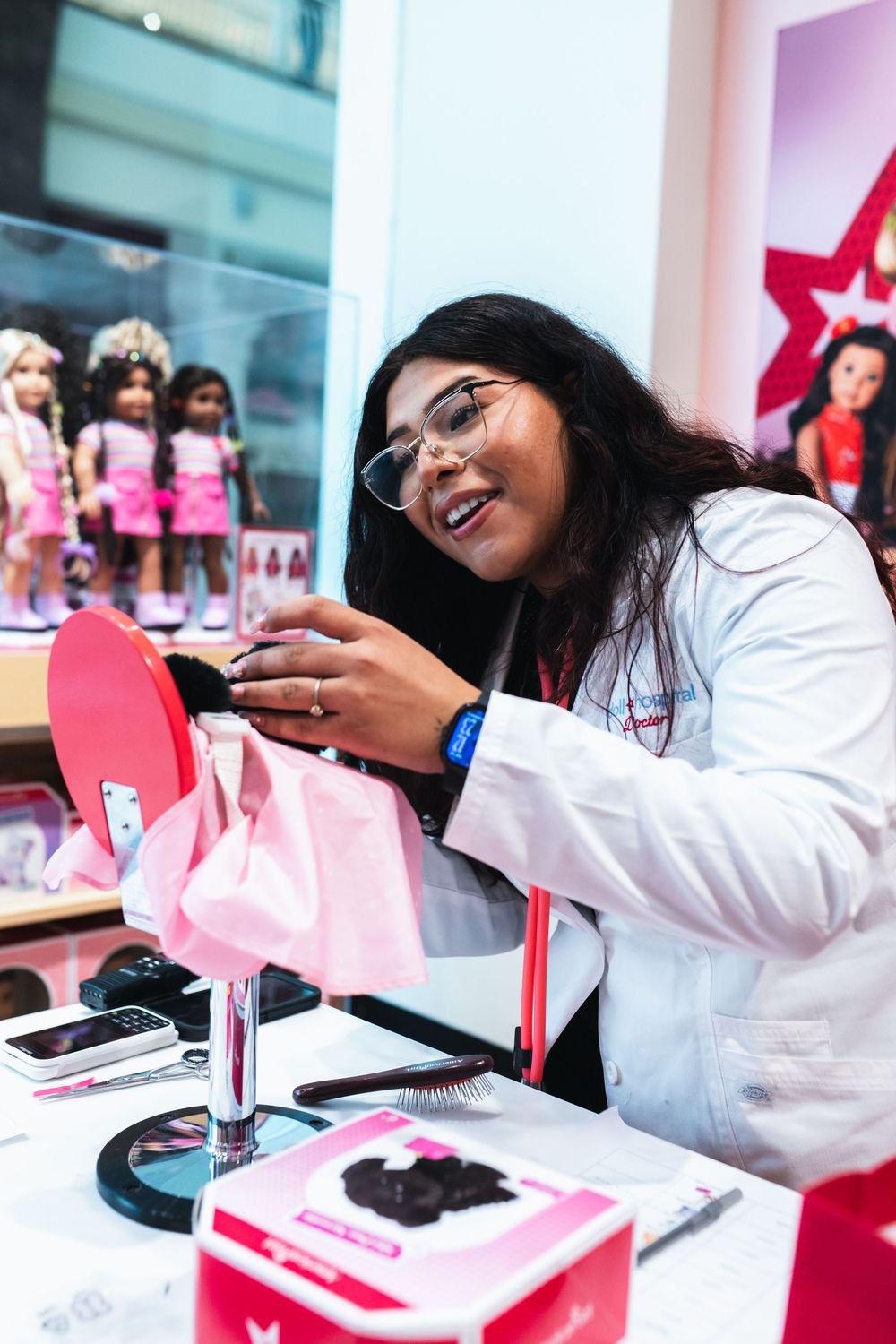 A smiling woman in a white lab coat styles a doll's hair at an American Girl store display.