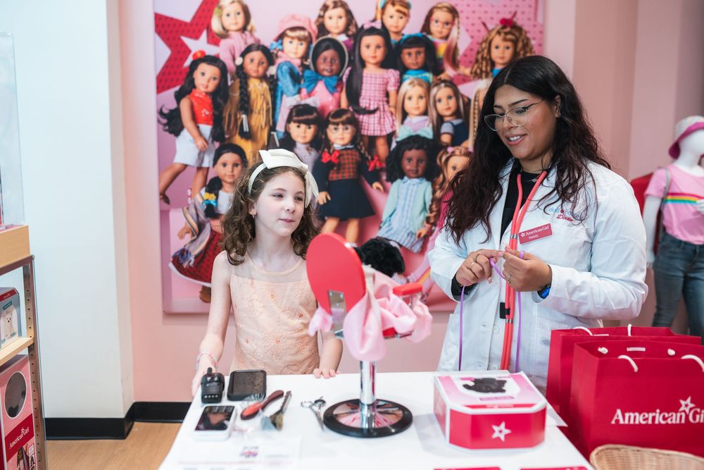 A girl and store associate explore American Girl products at a display table in a bright retail store.