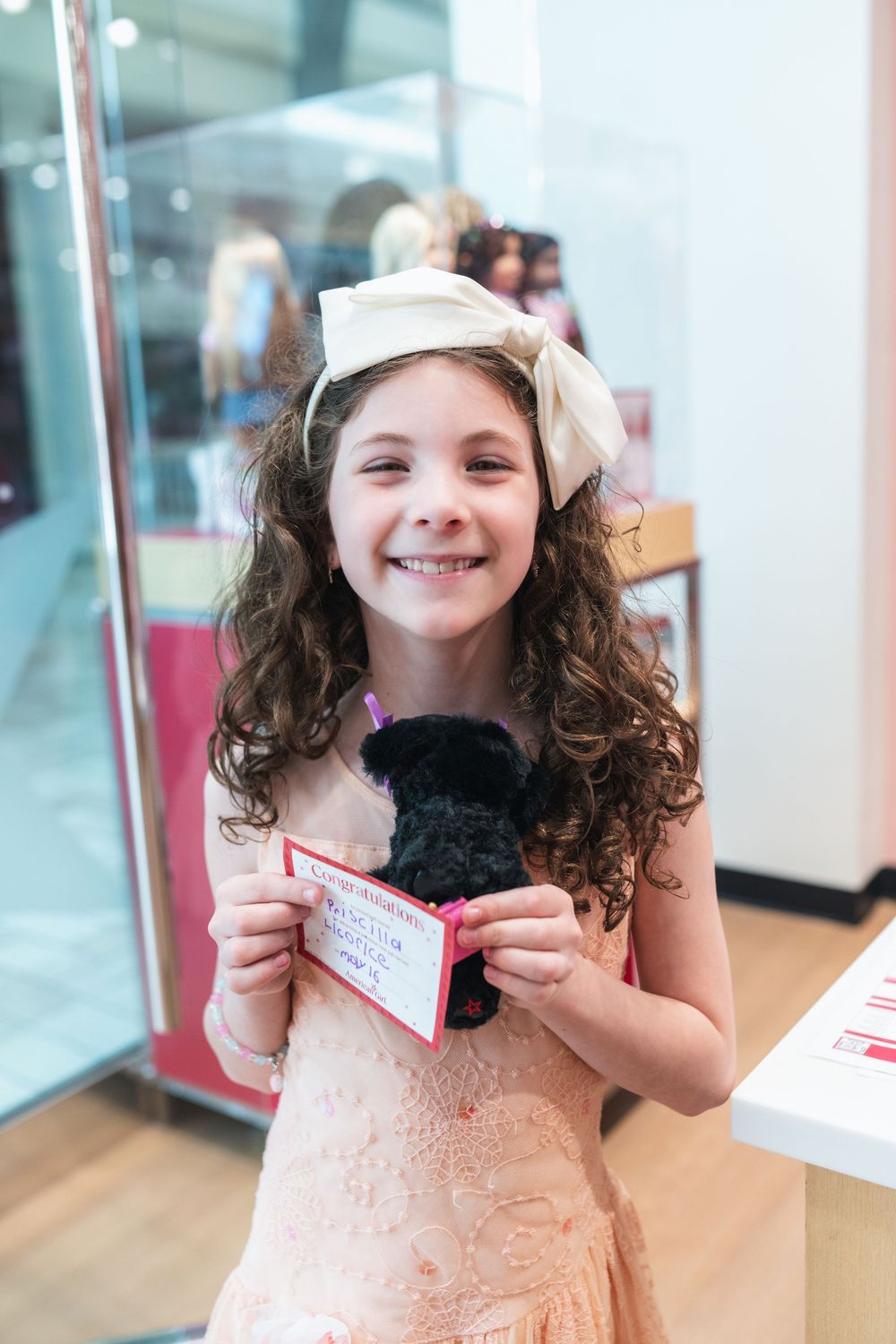 Smiling girl with curly hair and bow headband holding a small black stuffed animal and card in a store.