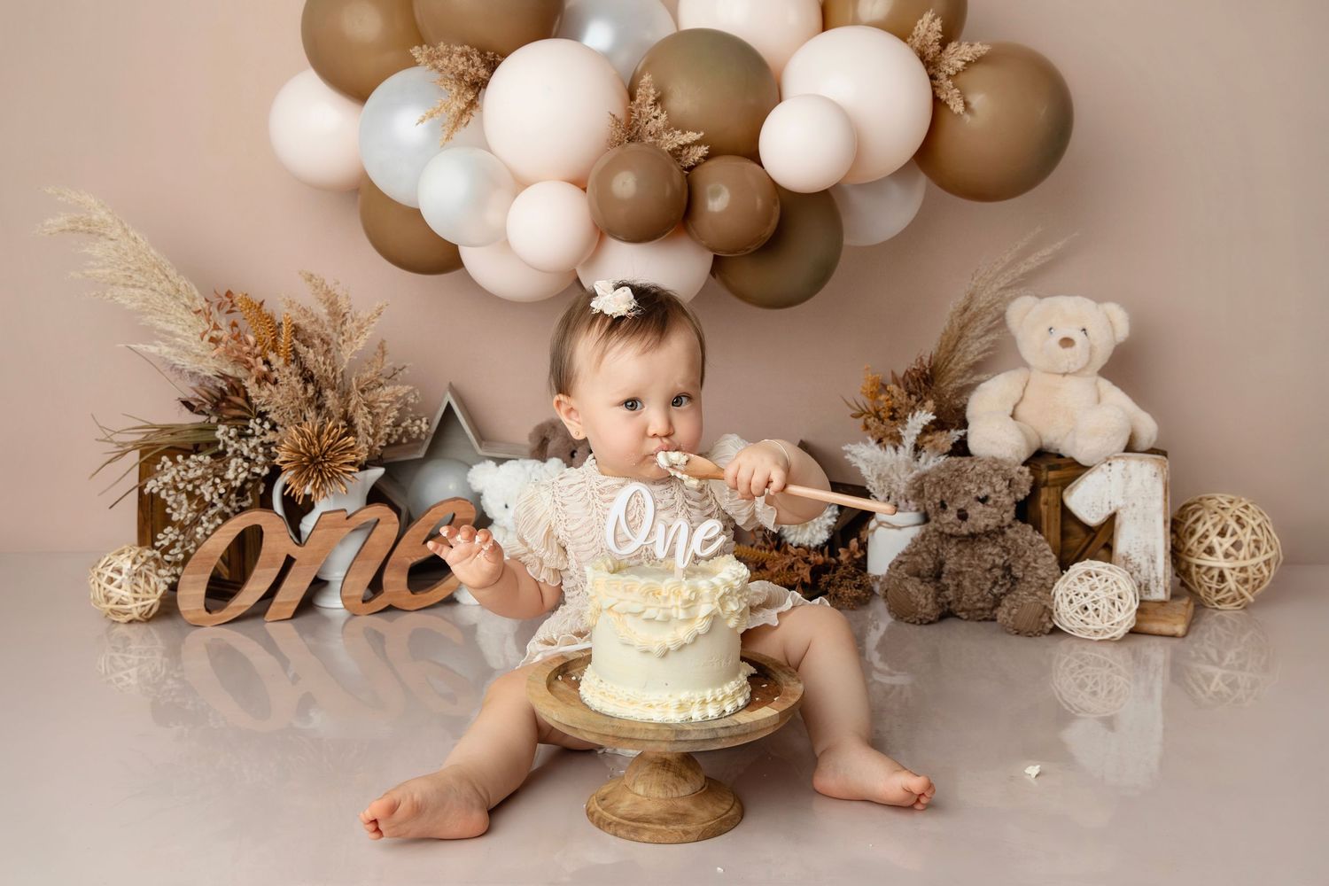 Baby girl celebrates first birthday smashing a white cake surrounded by teddy bears and balloon garland decorations.