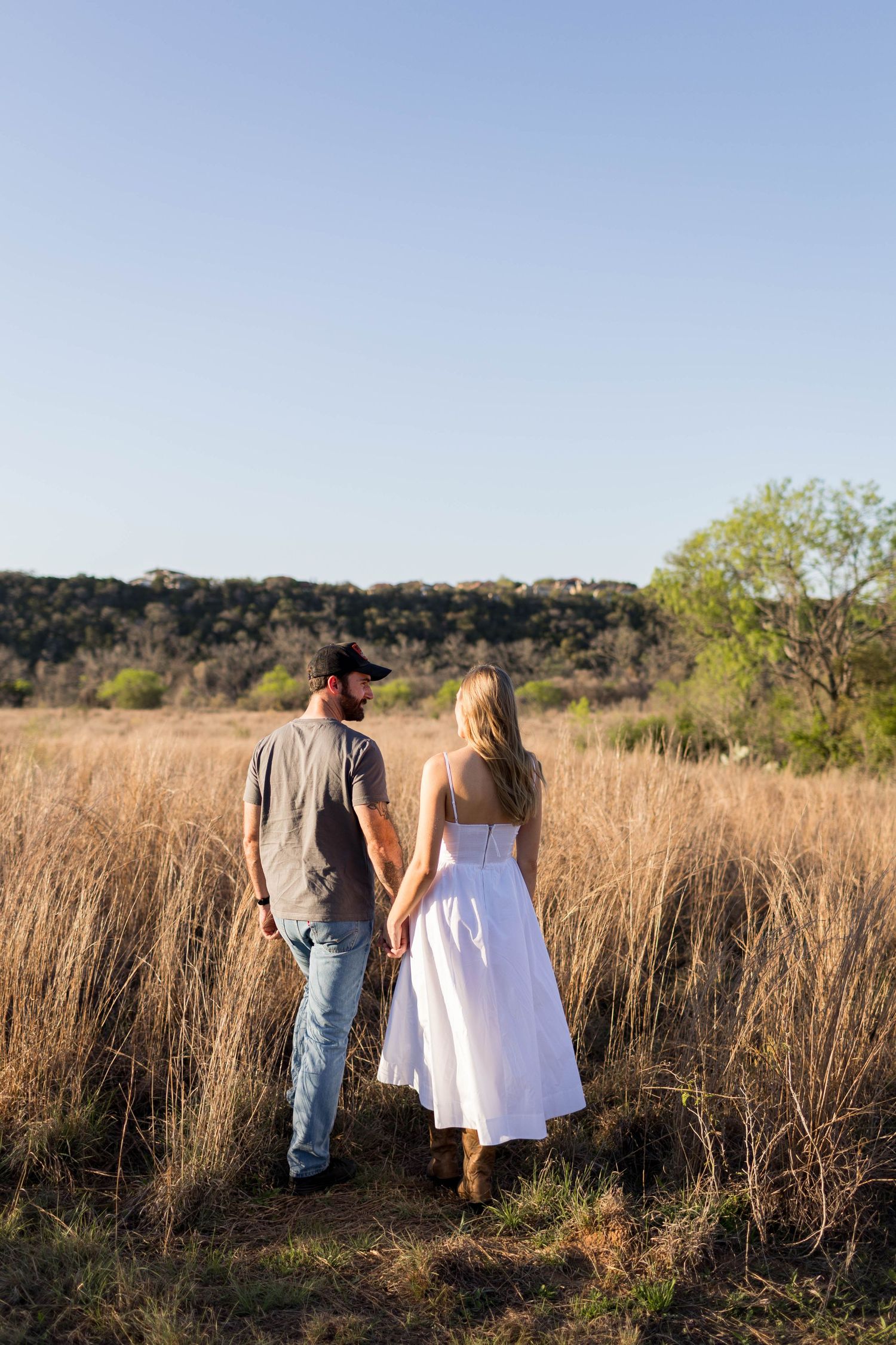A couple walks hand-in-hand through a golden field, the woman in a white dress and the man in jeans taken by Nicole Parker Photography.