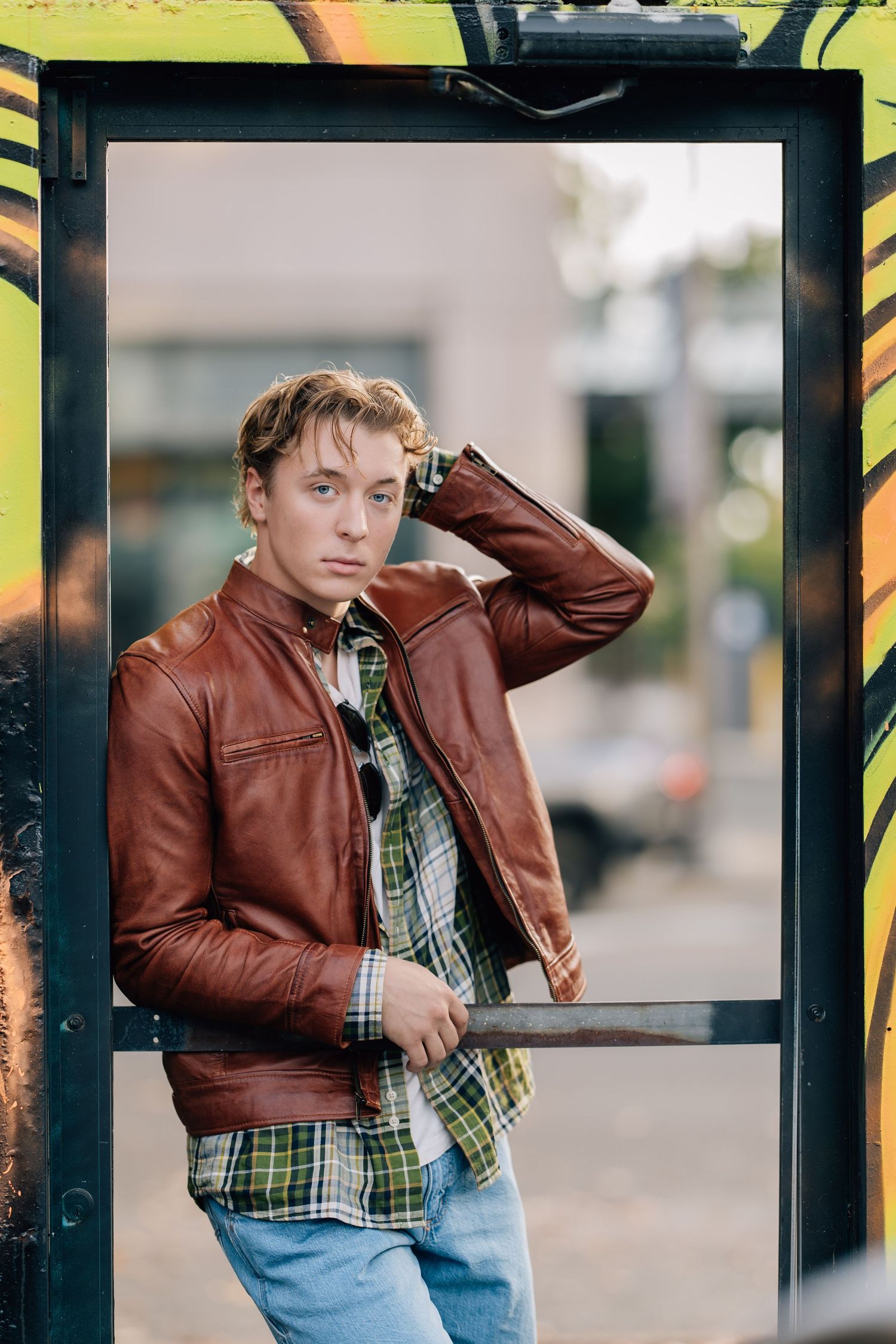 Young man in brown leather jacket and plaid shirt leaning against urban bus stop with colorful graffiti background.